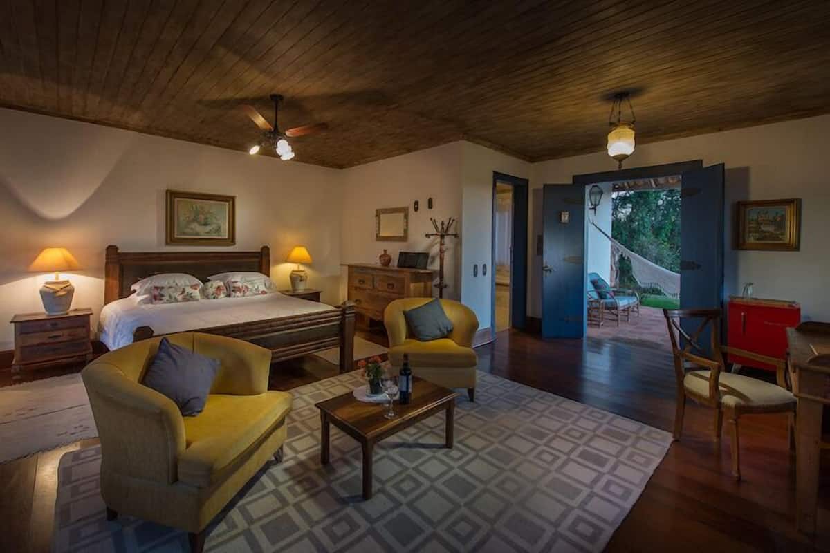 Una habitación acogedora con una cama, un sofá, una mesa de centro y un escritorio. Hay una puerta que da a un balcón con mobiliario exterior.