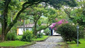 Uma passagem de paralelepípedos leva a um edifício com telhado em cerâmica, rodeado por vegetação exuberante e plantas floridas.