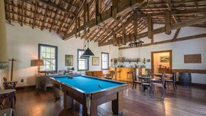Amplo quarto com mesa de bilhar, vigas de madeira no teto e um espaço bar com banquetas.