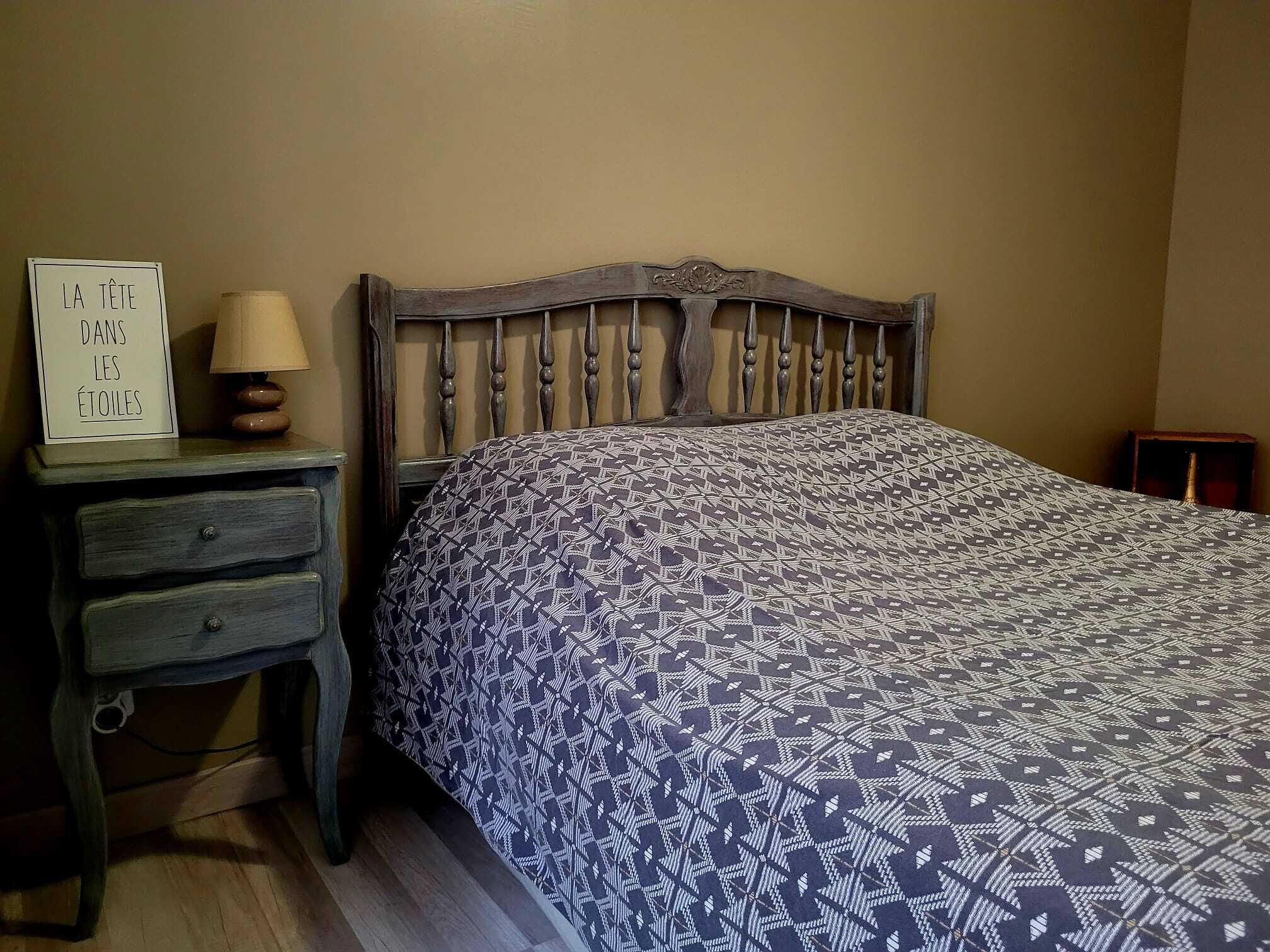 Un dormitorio con cabecera de madera, edredón estampado, mesita de noche con lámpara y un cartel en la pared.