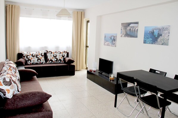 A modern living room with a sofa, TV, dining table, and chairs. There are three framed pictures on the wall.