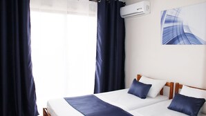 Hotellihuone, jossa on sänky, kaksi tyynyä, peitto, ikkuna verhoilla, ilmastointilaite ja seinälle kiinnitetty taideteos.