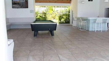 Una mesa de billar en una habitación con suelo de baldosas, una zona de bar con taburetes blancos y vistas a zonas verdes exteriores.