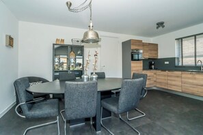 A modern dining area with a round table, four grey chairs, a decorative metal pendant light, and a kitchen with wooden cabinets and a black countertop.