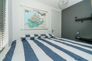 A bedroom with a bed, a map on the wall, a desk with a lamp, and a window with blinds.
