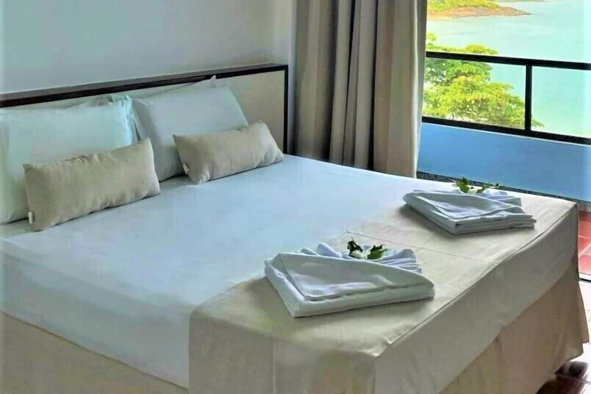 A neatly made bed with pillows and folded towels, a window with curtains, and a view of a landscape.