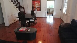 Een ruime woonkamer met houten vloeren, een eethoek met tafel en stoelen, een zwart voetenbankje met boeken, en een trap.