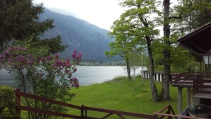 Vista al lago con muelle de madera, zona verde y montaña al fondo.