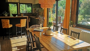 Un vaste espace dédié à la salle à manger, avec une longue table en bois et des chaises, un luminaire suspendu en rotin et de grandes fenêtres offrant une vue sur la verdure environnante.
