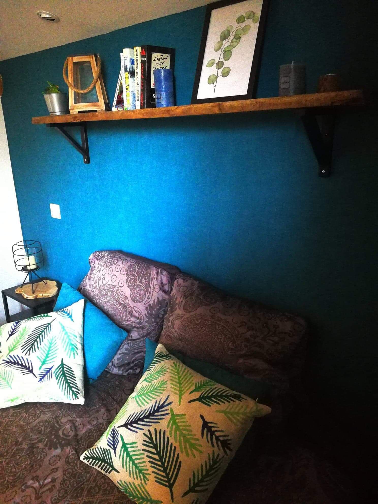En hyggelig stue med blå væg, en træhylde med bøger og dekorative genstande, en stribet sofa og to puder med bladmotiv.