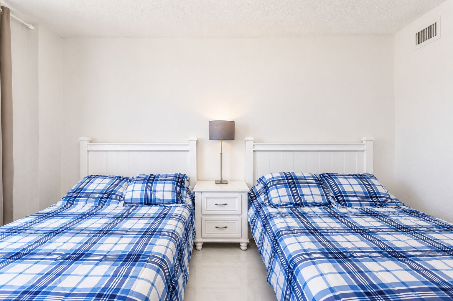 A twin room with two beds, each with a matching blue and white checkered bedspread, a bedside table with a lamp, and a white wall with a vent.