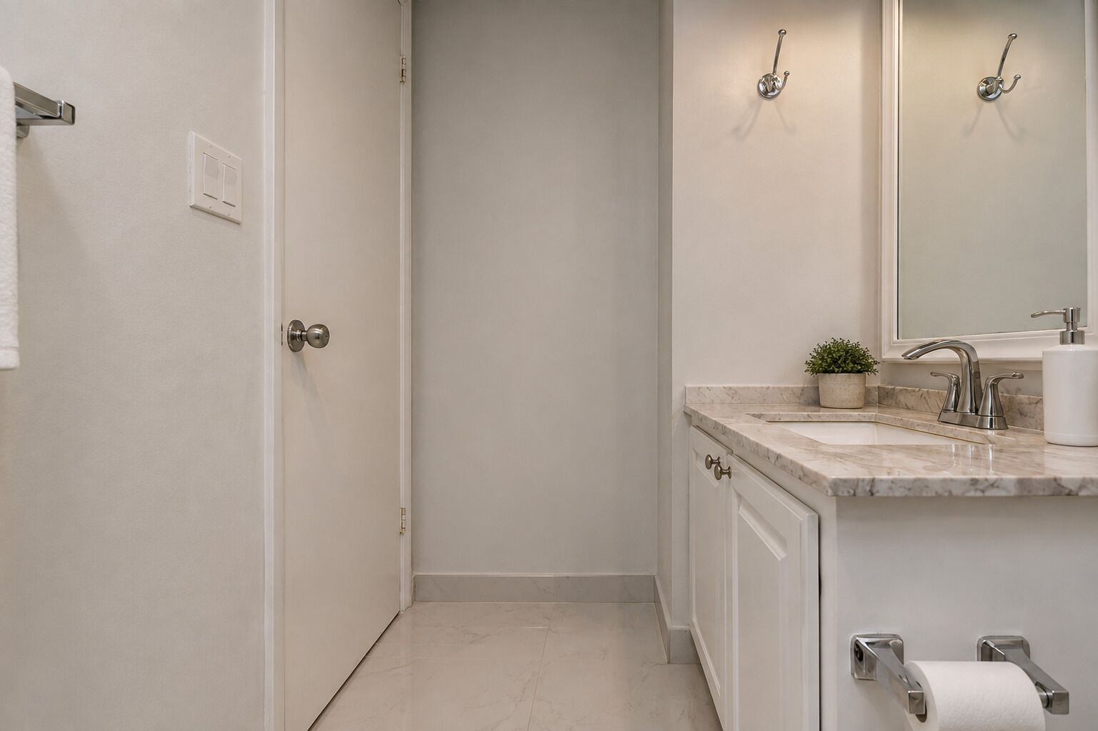 Une salle de bain avec une porte blanche, un lavabo doté d’un plan de travail en marbre, un miroir, une petite plante en pot et un porte-rouleau de papier toilette.