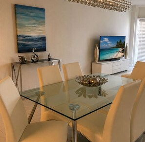 Ein moderner Essbereich mit einem Glastisch, weißen Stühlen, einer dekorativen Schale und einem Fernseher, der eine Strandansicht zeigt.