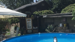 A clear blue swimming pool with a metal ladder, surrounded by a wooden deck and a house with a stone wall and a satellite dish. - Welcome to the "Hohe Liebe" vacation apartment (Bad Schandau-Ostrau)