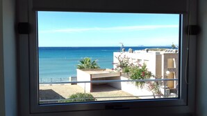 Blick aus dem Fenster auf eine Küstenregion mit blauem Himmel, ein Gebäude mit Balkon und ein Sandstrand.