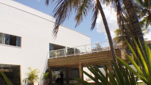 A modern building with a white facade, a covered outdoor area with seating, and palm trees. - Hotel Abrolhos (Nova Viçosa)