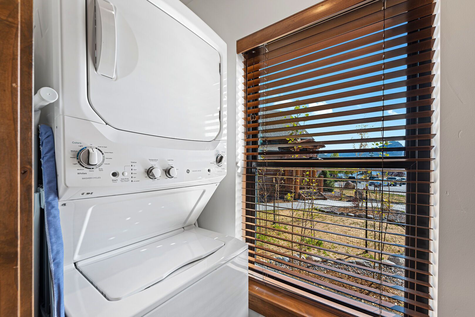 A front-loading washer and dryer unit with control knobs and a start button, positioned next to a window with horizontal blinds.