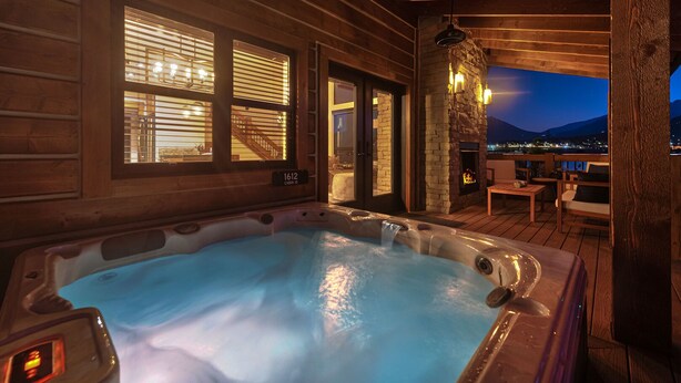 A hot tub in a wooden cabin with a stone fireplace, a view of the mountains, and a balcony with outdoor seating.