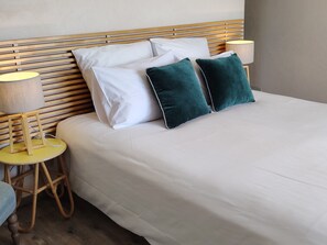 Una cama bien hecha con sábanas blancas y dos almohadas verdes, flanqueada por dos mesitas de noche con lámparas, una de ellas con estructura de madera.