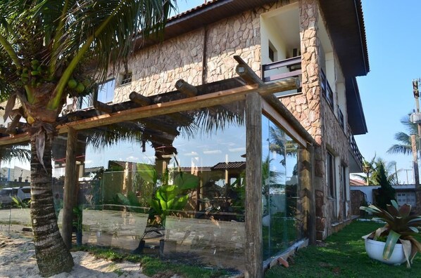 Un edificio de piedra con un balcón, una pérgola con cerramiento de vidrio y una palmera.