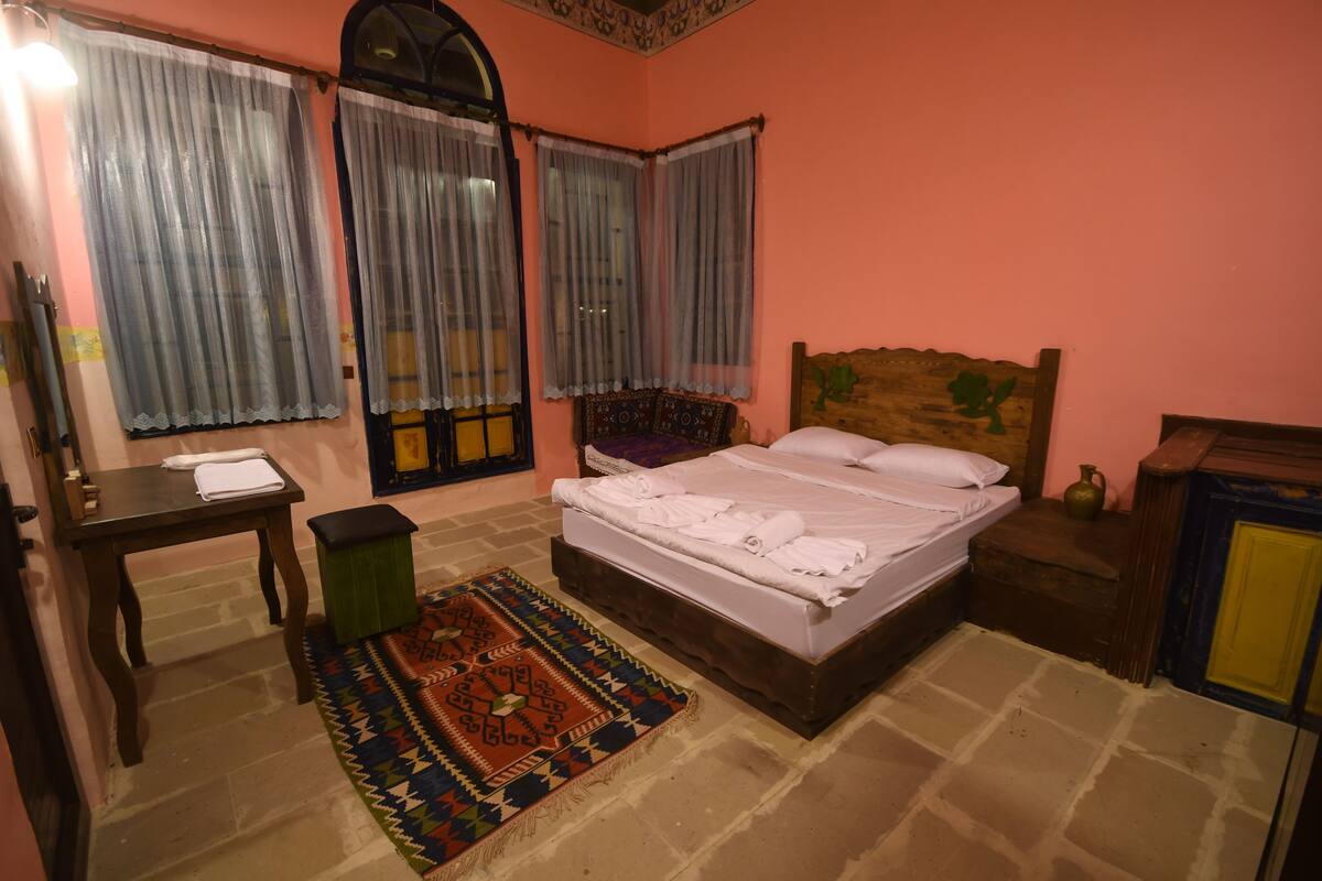 Quarto com cama, cabeceira de madeira, mesa de cabeceira, mesa, banco e tapete estampado.