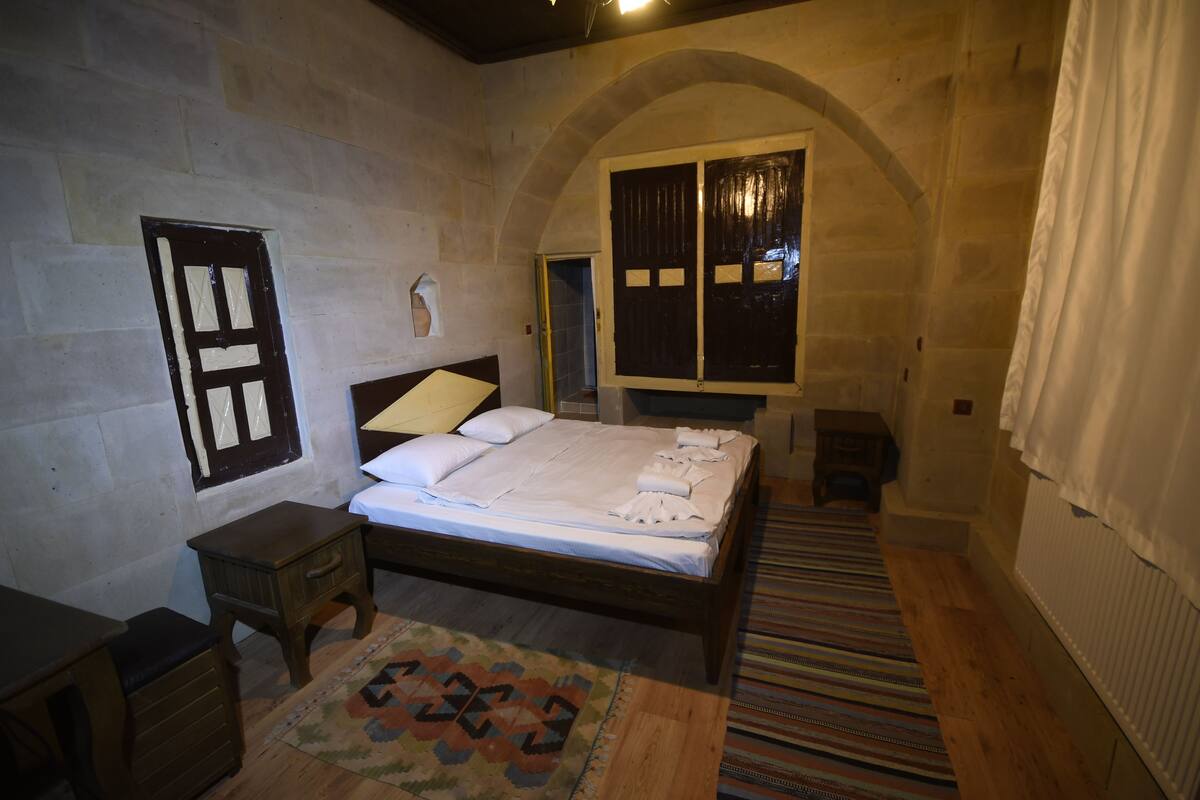 Quarto com cama de madeira, mesa de cabeceira, pequena mesa, tapete estampado e uma porta arqueada.