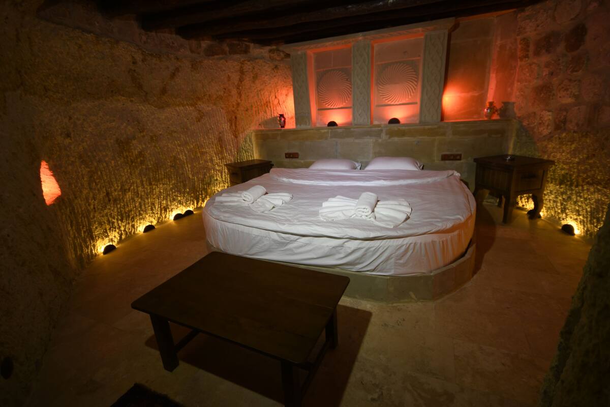 Quarto com pouca luz, parede de pedra, cama grande com roupa de cama branca e um banco de madeira.