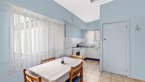 A compact kitchen with white cabinets, a stove, and a sink. There is a dining table with chairs and a window with patterned curtains. - Hutton Hide Away - East Coast Getaways (The Entrance North)