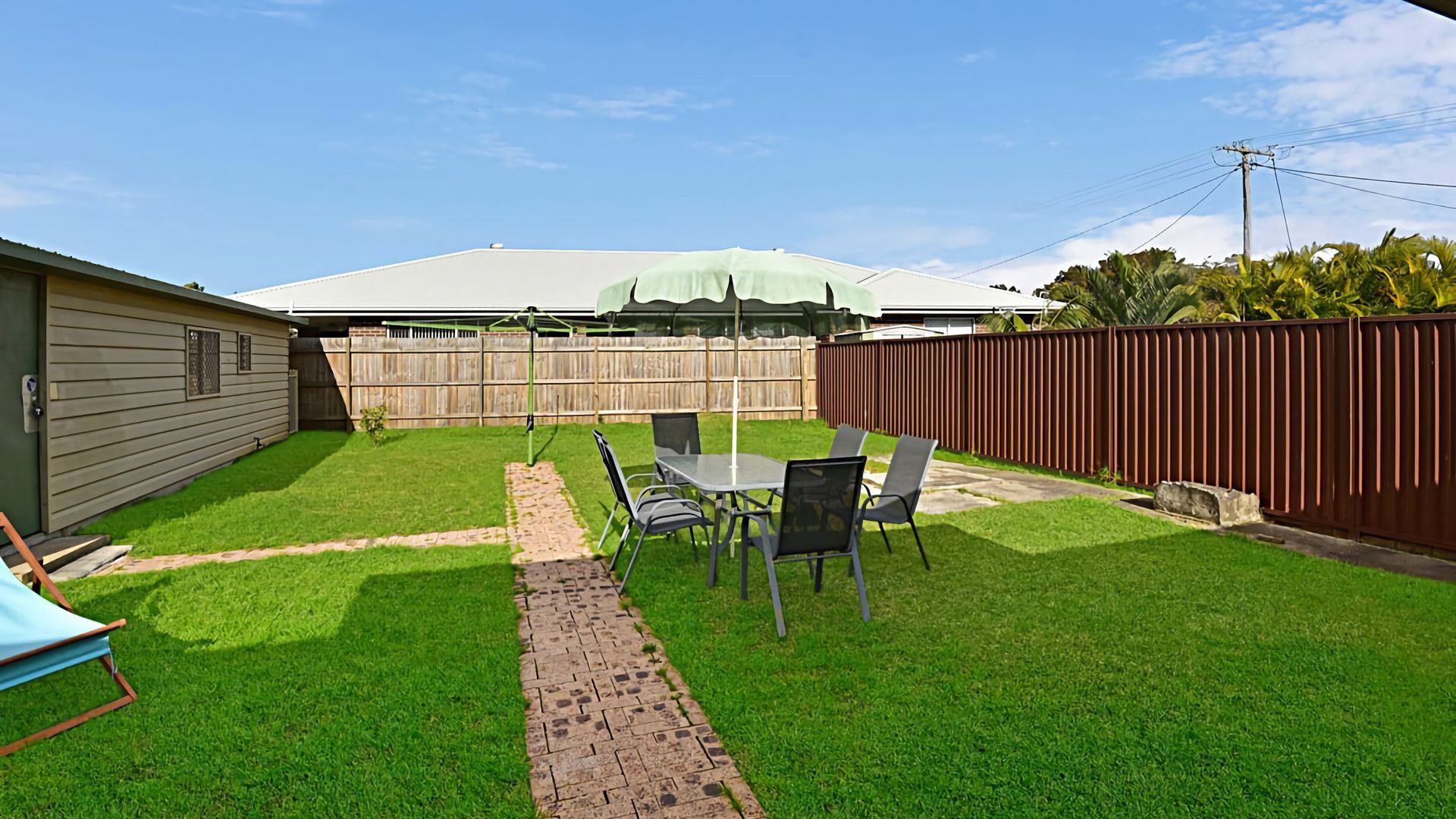 A backyard with a patio table and chairs, a brick pathway, a wooden fence, and a house with a green roof.