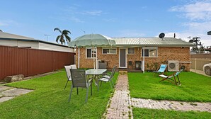 Ein Garten mit einem Ziegelhaus, einem Patio-Tisch mit Stühlen, einer Rasenfläche und einem überdachten Bereich.