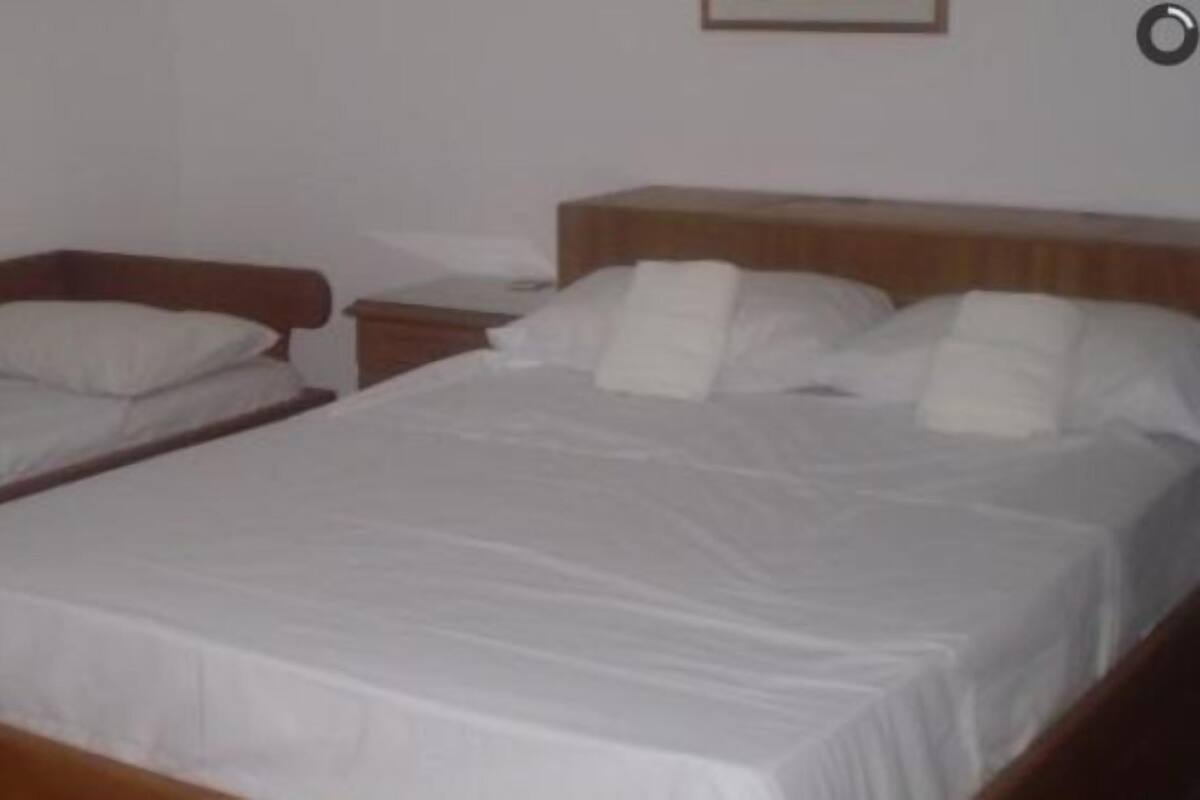 A room with two single beds and one double bed, wooden headboards, white bedding, a nightstand, and a window with curtains.
