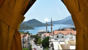 A view through a window with curtains, featuring a street, buildings, a mosque with two minarets, a dome, and a body of water with mountains in the background. - Limon Pansiyon (Kas)