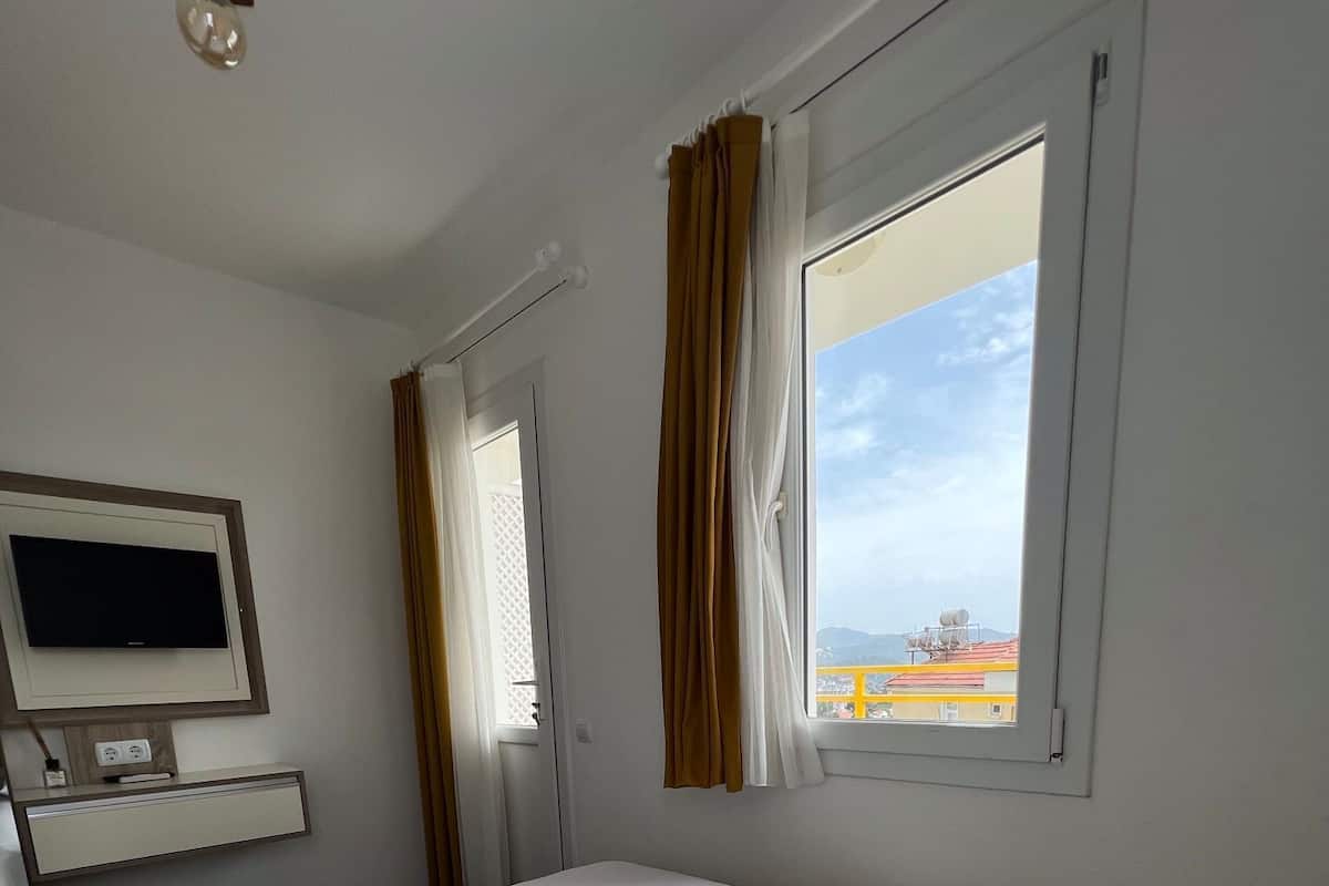 A hotel room with a large bed, a flat-screen TV mounted on the wall, a window with curtains, and a view of the cityscape.