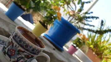 Une tasse de café sur une table, accompagnée d’une assiette décorative, le tout entouré de plantes en pot et, en arrière-plan, d’un bâtiment sur lequel on peut voir une enseigne.