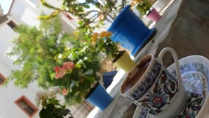 A cup of coffee on a saucer with a floral design, placed on a table outdoors with potted plants and a building in the background. - Limon Pansiyon (Kas)