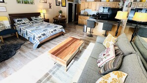 Een moderne woonkamer met een groot bed, een houten salontafel en een grijze bank met gekleurde kussens. Er is een keukenhoek met houten kasten en een eettafel.