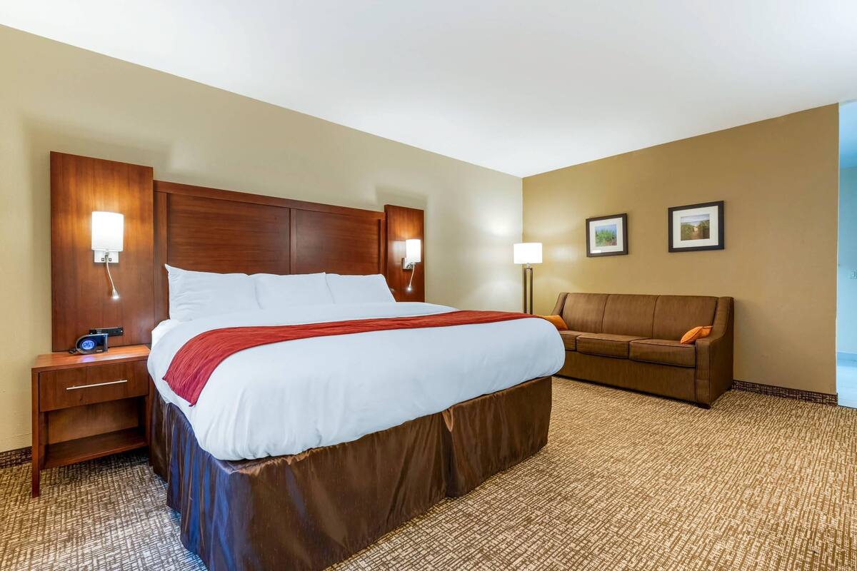 A hotel room with a large bed, a wooden headboard, a nightstand with a lamp, a brown sofa, and a carpeted floor.