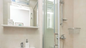 Un baño moderno con ducha envuelta en vidrio, un lavabo blanco con espejo y un pequeño estante con artículos de aseo.