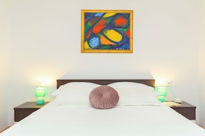 Cama bem arrumada com uma almofada decorativa, duas luminárias de cabeceira e um quadro abstrato colorido na parede.