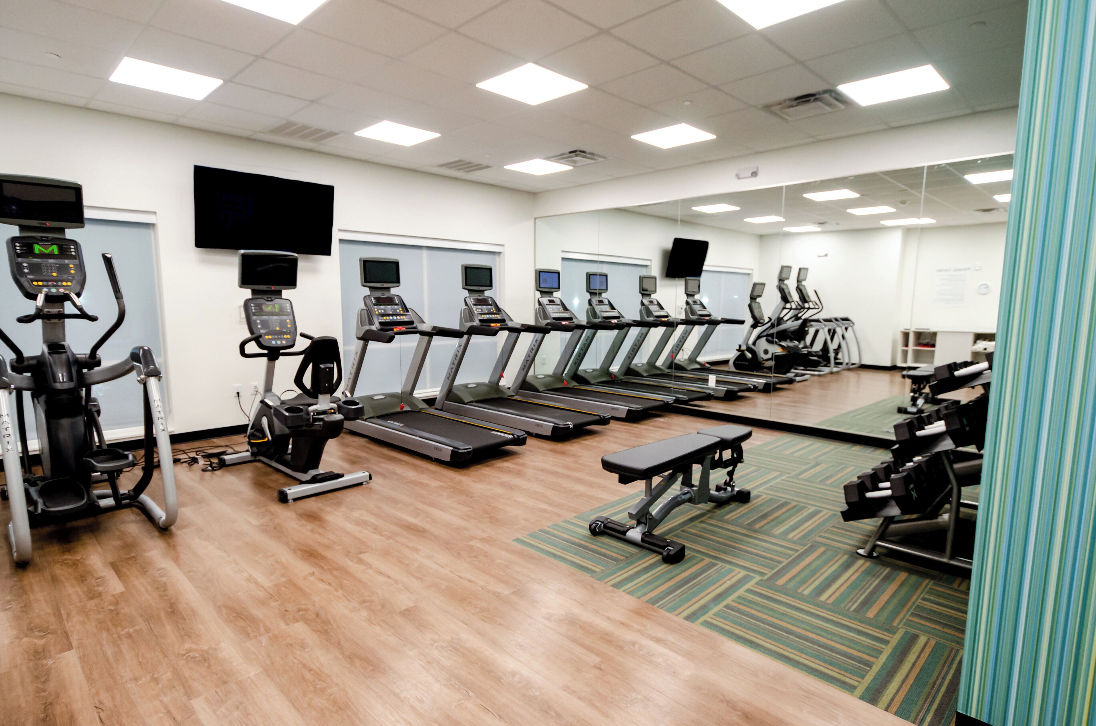 A gym with multiple treadmills and exercise bikes, a wooden floor, and a large mirror wall.