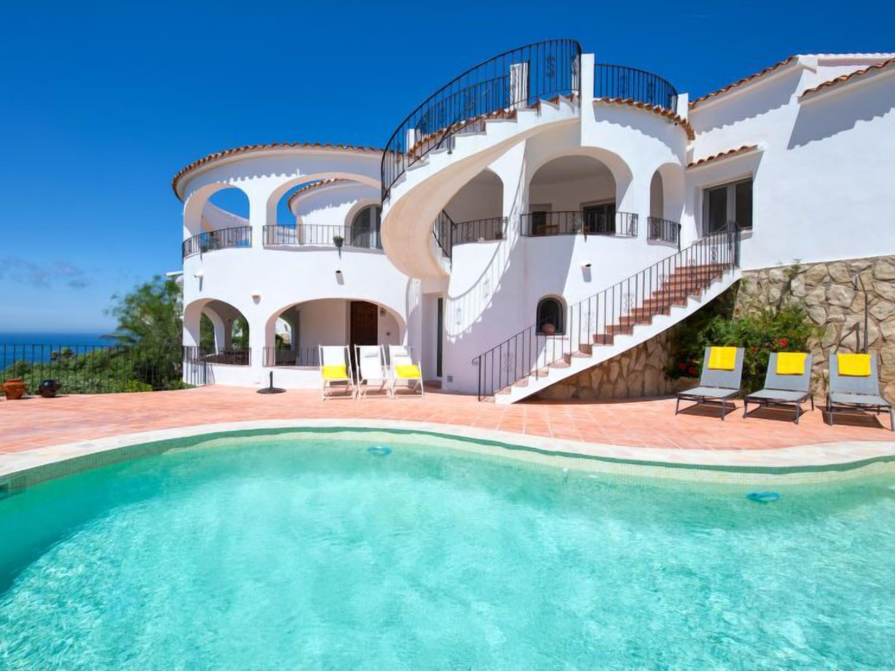 A multi-story white building with a curved swimming pool, outdoor seating, and a view of the ocean.
