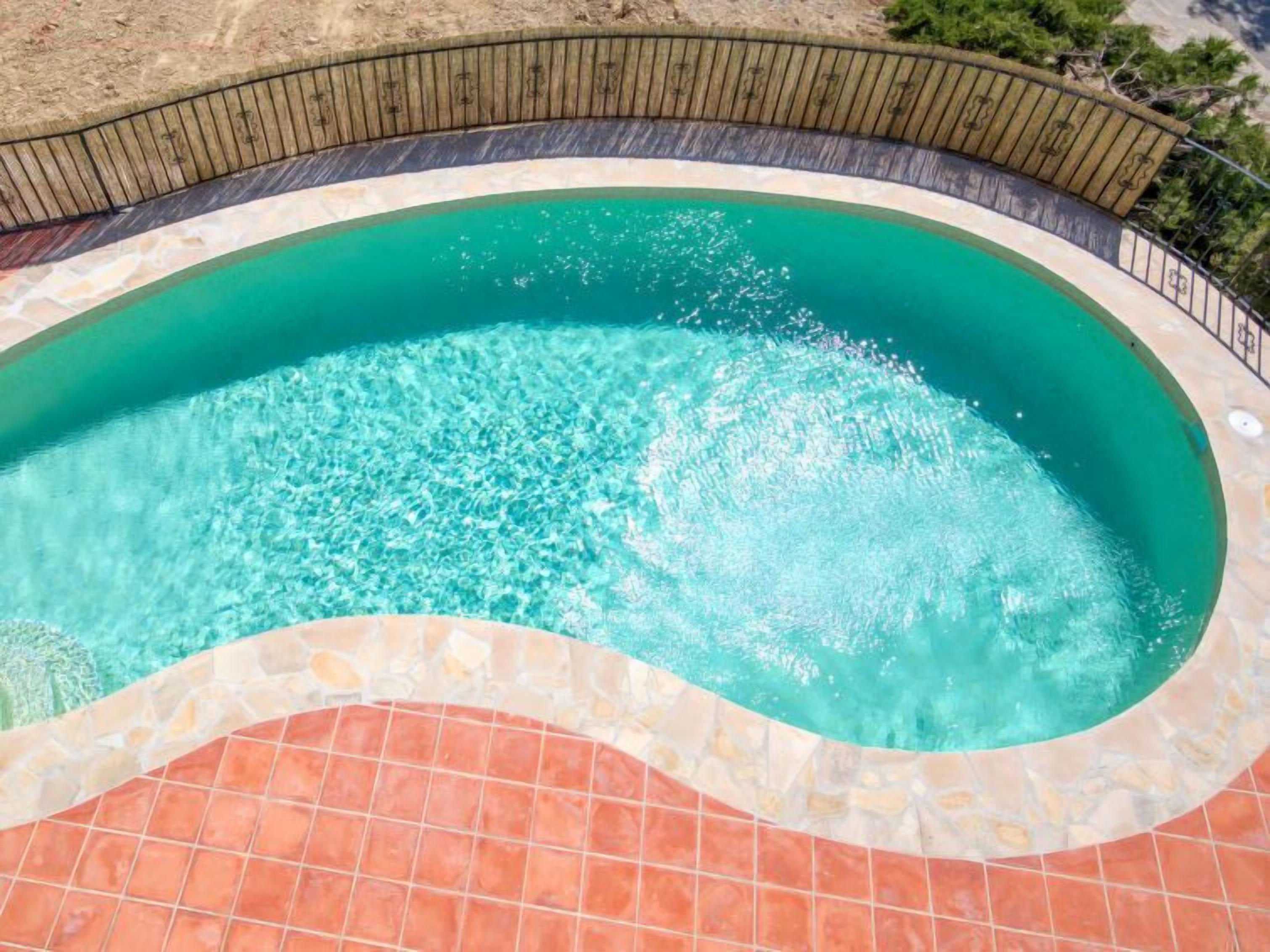 A swimming pool with clear water, surrounded by a stone and tile border, and a wooden fence.