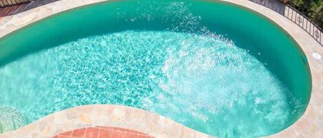 A swimming pool with clear water, surrounded by a stone and tile border, and a wooden fence.