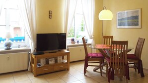 A living room with a TV on a wooden stand, a dining table with chairs, and a painting on the wall. - bright terrace apartment with beach chair in the old captain's house, max. 3 pers. (Prerow)
