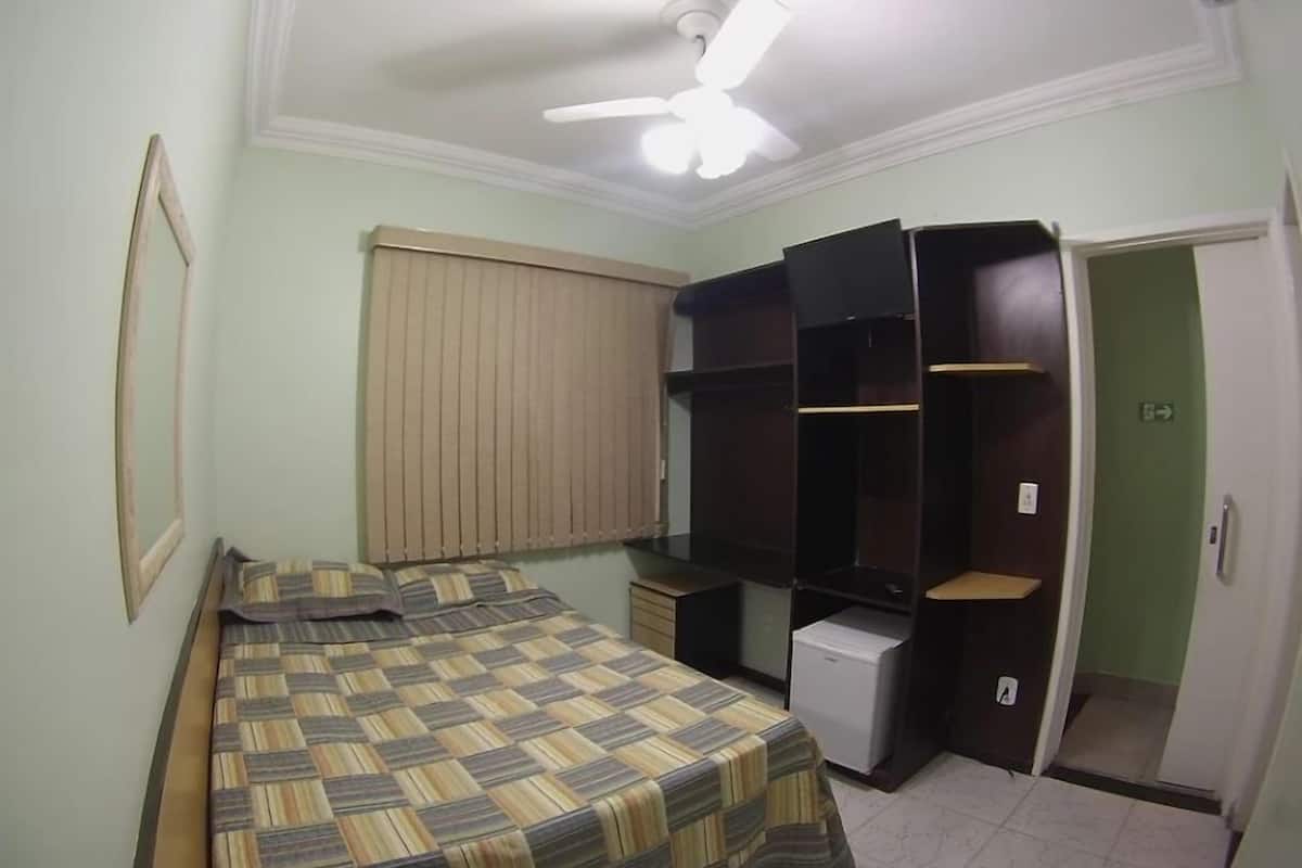 Un dormitorio con cama, ventilador de techo, ventana con persianas, armario y un refrigerador pequeño.