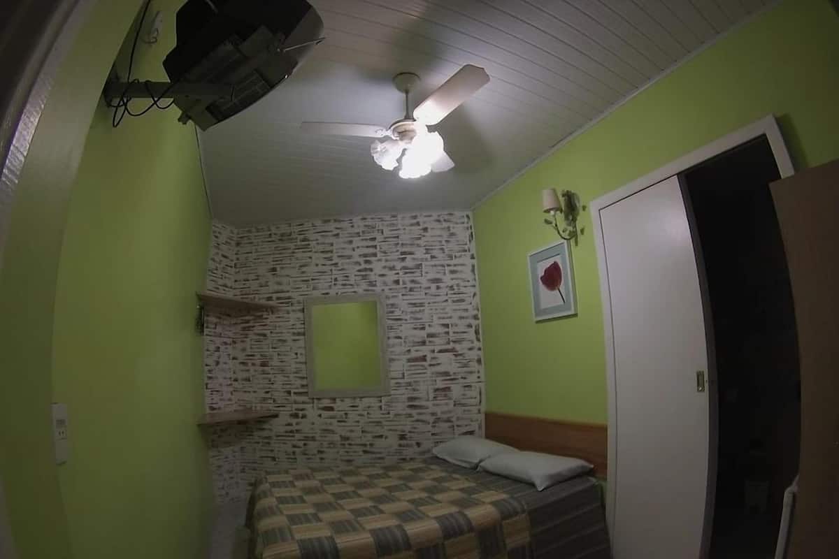 Una habitación con una cama, un ventilador de techo, un estante en la pared, un cuadro enmarcado y una puerta.