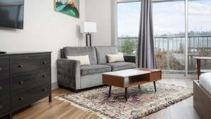 A modern hotel room with a grey sofa, a wooden coffee table, a patterned rug, a flat-screen TV, and a painting on the wall. - Studio King Suite with Kitchen | 203 (Asheville)