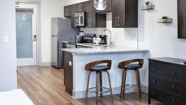A modern kitchen with dark cabinets, stainless steel appliances, a granite countertop, and two wooden stools. - Studio King Suite with Kitchen | 203 (Asheville)