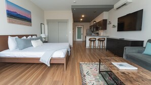 A modern hotel room with a bed, a sofa, a coffee table, and a kitchen area with a TV. - Studio King Suite with Kitchen | 203 (Asheville)