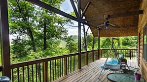 Um deck de madeira com uma cadeira de balanço e uma mesa, com vista para uma área arborizada.
